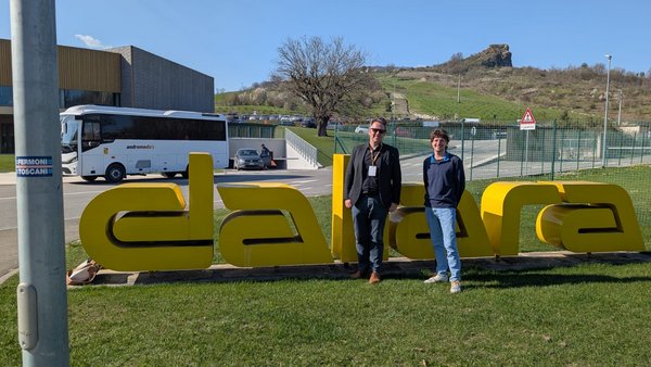 Zwei Männer stehen nebeneinander vor einem großen Schriftzug „dallara“ auf einer Wiese